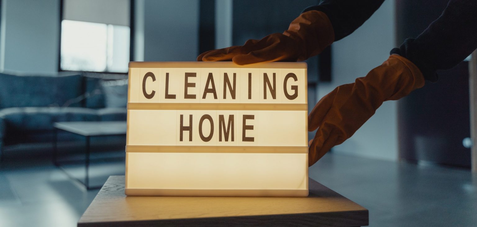 Banner dla artykułu na temat ozonowania - na obrazku widać tabliczkę z napisem cleaning home stawianą na stole przez osobę w pomarańczowych rękawiczkach
